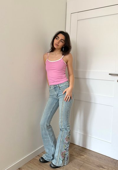 Joven mujer con camiseta sin mangas rosa y jeans azules con bordado floral apoyada contra una pared cerca de una puerta blanca cerrada, mirando de lado.