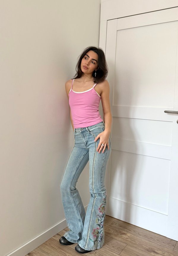 Giovane donna con canottiera rosa e jeans blu con ricamo floreale appoggiata al muro vicino a una porta bianca chiusa, guarda di lato.