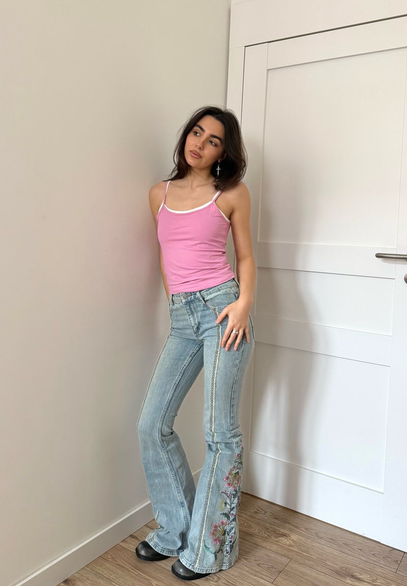 Νεαρή γυναίκα με ροζ τοπ χωρίς μανίκια και μπλε τζιν με κεντήματα λουλουδιών στηρίζεται σε τοίχο κοντά σε κλειστή λευκή πόρτα, κοιτάζοντας πλάγια.