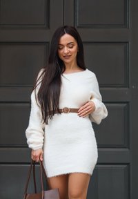 Femme aux longs cheveux bruns portant une robe blanche duveteuse avec une ceinture marron, tenant un sac cabas marron, debout devant des portes à panneaux foncées.