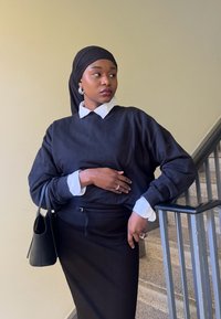 Schwarzer Pullover mit weißem Shirt darunter, kombiniert mit einem schwarzen Rock. Die Accessoires umfassen Creolen und eine schwarze Handtasche.