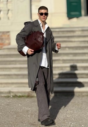 Grauem Trenchcoat, weißem Hemd, dunkelgraueren Hosen, schwarzen Schuhen und brauner Lederhandtasche. Sonnenbrille vervollständigt den Look, im Freien mit Treppen im Hintergrund.