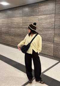 Surchemise jaune clair, sac bandoulière noir, pantalon noir ample et bonnet à motifs avec des rayures marron, noir et crème. Fond de mur lisse.