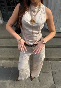 Haut beige sans manches et pantalon large en tissu texturé, accessoirisés avec plusieurs colliers en or, bagues et une ceinture à motifs.