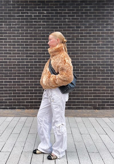 Chaqueta de piel sintética en marrón claro con manchas blancas, cuello alto, combinada con pantalones cargo blancos y bolsa negra. Los zapatos tienen un diseño de estampado de serpiente.