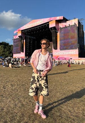 Joven con camisa rosa y pantalones cortos con estampado de vaca posa en un festival al aire libre con un gran escenario y una multitud al fondo.