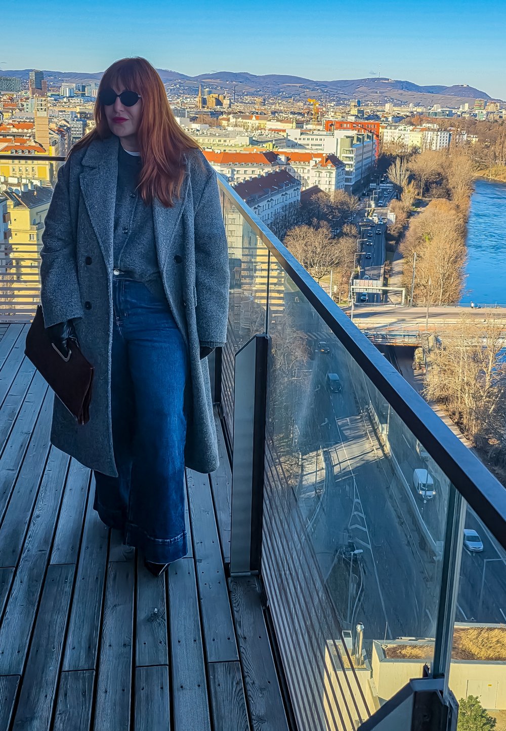 Kobieta w szarym płaszczu i okularach przeciwsłonecznych stoi na balkonie na dachu, z widokiem na panoramę miasta z rzeką i wzgórzami w słoneczną pogodę.