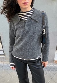 Γυναίκα που φοράει γκρι μάλλινο πουλόβερ πάνω από ζιβάγκο με ασπρόμαυρες ρίγες, μαύρο δερμάτινο παντελόνι και κρατάει μια μικρή μαύρη τσάντα ώμου.