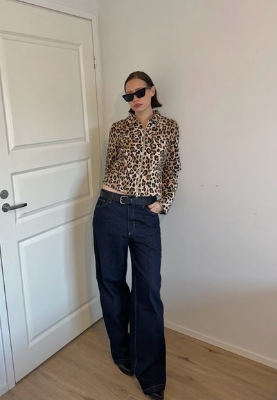Chaqueta con estampado de leopardo y cremallera, con mangas largas, combinada con pantalones vaqueros de pierna ancha en azul oscuro, con costuras en contraste, y zapatos de punta negra.