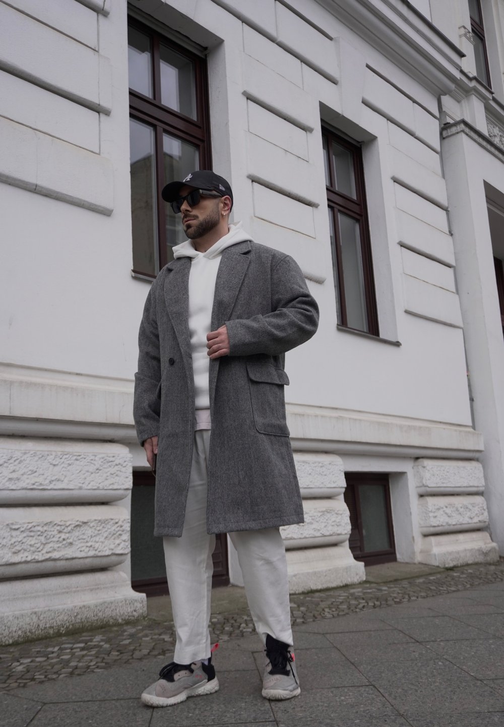 Casaco de lã cinza com colarinho em bico, sobre um hoodie branco. Combinado com calças brancas e ténis cinzentos. Usando um boné preto e óculos de sol.