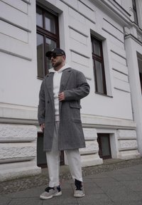 Casaco de lã cinza com colarinho em bico, sobre um hoodie branco. Combinado com calças brancas e ténis cinzentos. Usando um boné preto e óculos de sol.