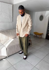 Μπεζ σακάκι πάνω από λευκό πουκάμισο, πράσινα χαλαρά παντελόνια και καφέ παπούτσια με λευκές λεπτομέρειες. Το ηλιακό φως ρίχνει σκιές στο πλακάκι του δαπέδου.