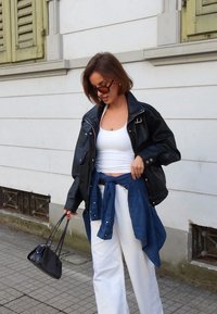 Veste en cuir noire portée sur un débardeur blanc, chemise en denim bleu nouée à la taille, et pantalon blanc à jambes larges. Petit sac à main noir. Cadre extérieur.