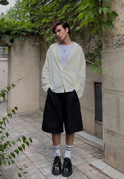 Camisa de botón amarilla clara sobre una camiseta blanca, pantalones cortos negros hasta la rodilla, calcetines blancos con rayas azules y zapatos negros con suela gruesa.