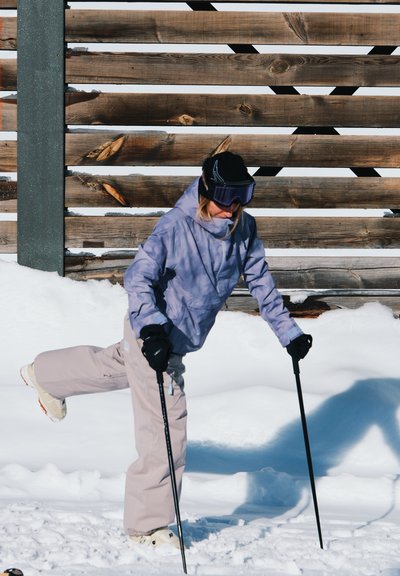 Persona esquiando, vestida con chaqueta lila clara, pantalones beige, guantes negros y gafas de esquí, usando bastones en nieve fresca con un fondo de madera.