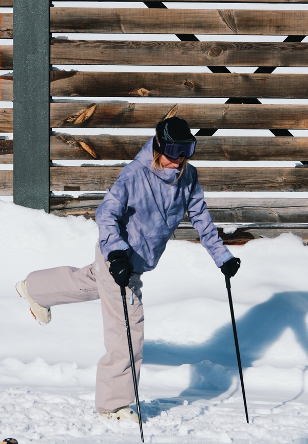 Skifahrende Person mit einer hellvioletten Jacke, beigen Hosen, schwarzen Handschuhen und Skibrille, die Stöcke im frischen Schnee vor einem hölzernen Hintergrund benutzt.