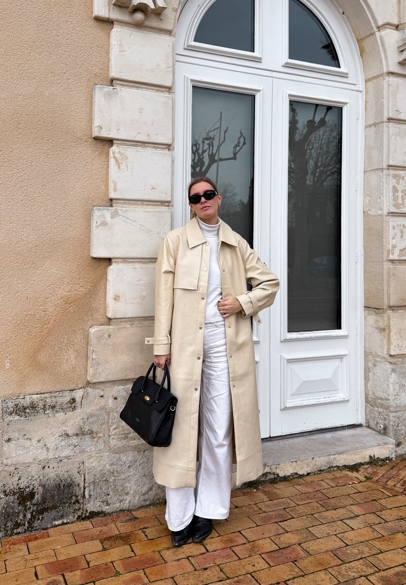 Femme en manteau long beige, col roulé blanc et pantalon, tenant un sac à main noir, portant des lunettes de soleil noires, debout près d'une porte blanche en arc sur un bâtiment en pierre.