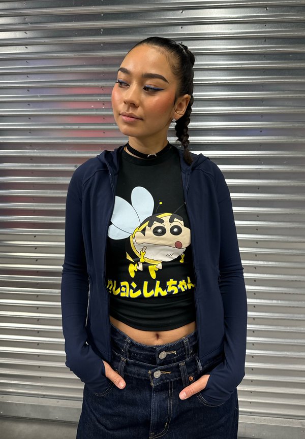 Jeune femme aux cheveux tressés portant une veste sombre et un t-shirt noir avec un dessin animé d'abeille et du texte japonais, debout devant un rideau métallique.