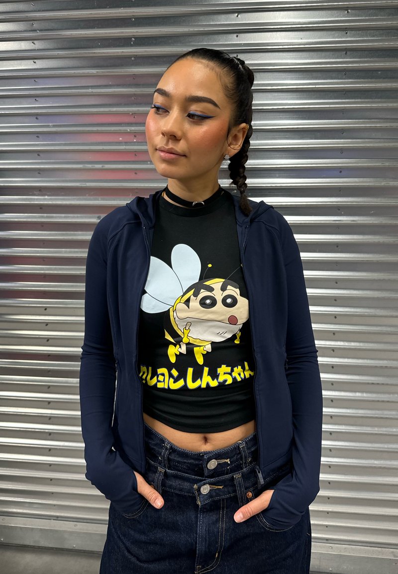 Jeune femme aux cheveux tressés, portant une veste sombre et un t-shirt noir avec un dessin animé d'abeille et du texte japonais, debout devant un volet métalique.