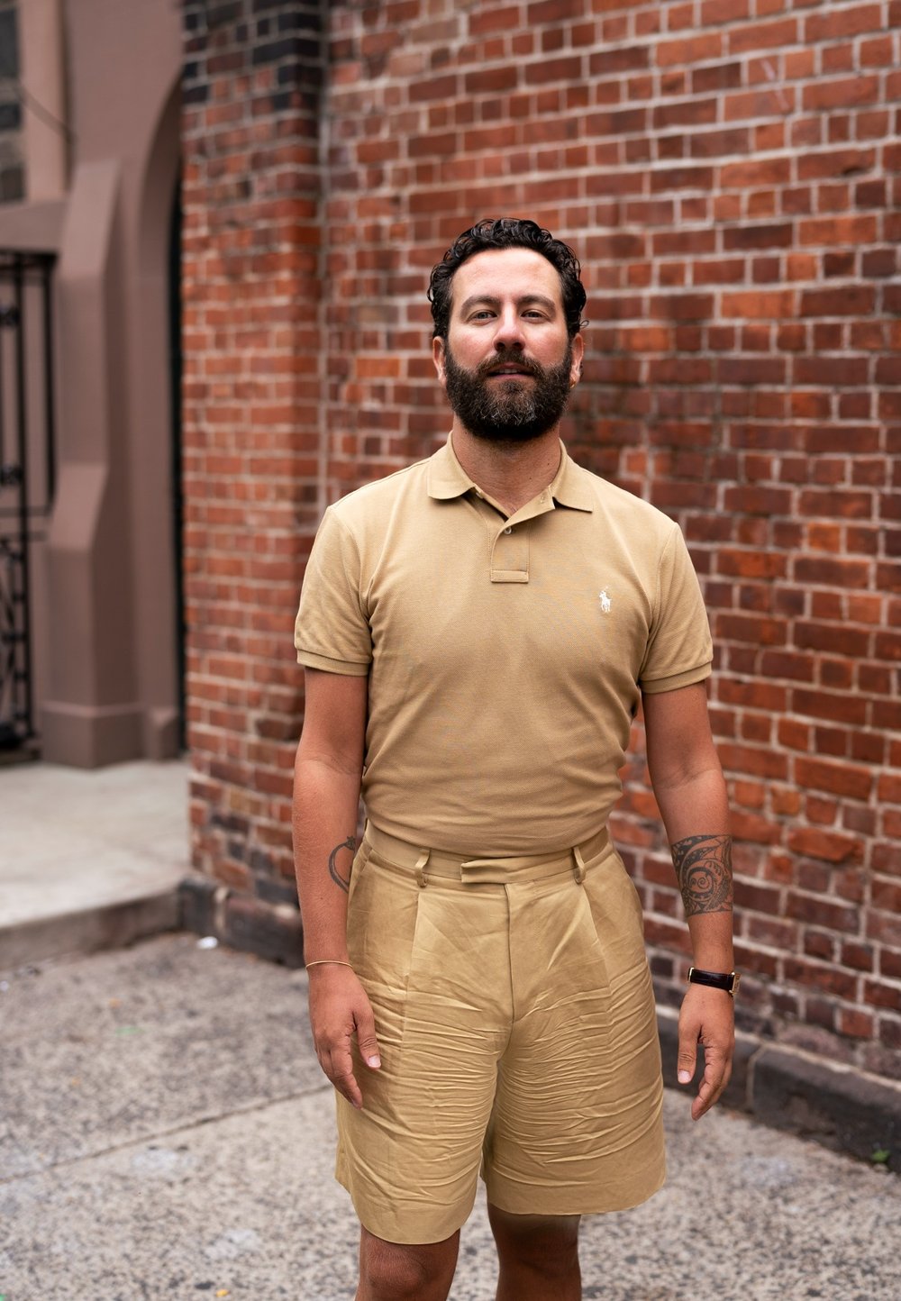 Ljusbrun polo t-shirt och matchande högmidjade shorts i slätt tyg, med subtila texturer. Bakgrunden har en röd tegelvägg.