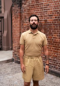 Ljusbrun polo t-shirt och matchande högmidjade shorts i slätt tyg, med subtila texturer. Bakgrunden har en röd tegelvägg.