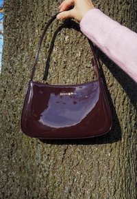 Bordeaux värinen patentoitu nahkalaukku, jossa on kaareva muoto ja yksi hihna. Varustettu kultaisilla tarvikkeilla, joissa on "kazar"-logo, ja sileällä kiiltävällä pinnalla.