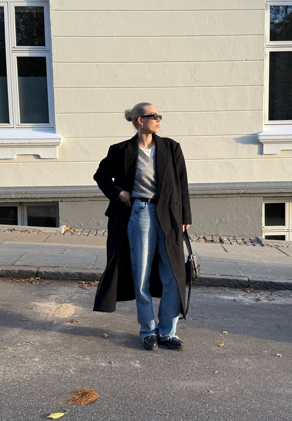 Μαύρο μακρύ παλτό από μάλλινη ύφανση πάνω από γκρι πουλόβερ, σε συνδυασμό με ανοιχτό μπλε φαρδύ τζιν και μαύρα παπούτσια. Αξεσουάρ με γυαλιά ηλίου και μαύρη τσάντα.