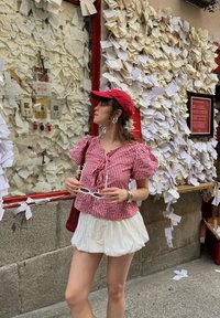 Czerwona i biała koszula w kratkę z bufkowymi rękawami oraz wiązaniem z przodu, w połączeniu z białą rozkloszowaną spódnicą. Dodatki obejmują okulary oraz czerwoną czapkę.