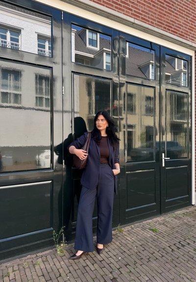 Blazer azul marino y pantalones de pierna ancha combinados con una blusa marrón. Sostiene un bolso texturizado marrón, de pie frente a una puerta verde con reflejos.