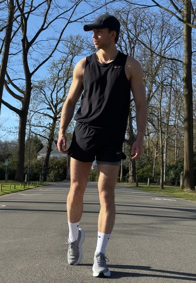 Camiseta deportiva sin mangas negra y pantalones cortos, zapatillas gris con detalles blancos y calcetines blancos. Entorno al aire libre con árboles y cielo azul.