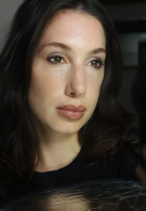 Donna con lunghi capelli scuri che guarda leggermente di lato con un trucco soft, labbra neutre e un'espressione calma in luce soffusa.