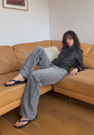 Mujer con cabello rizado recostada en un sofá de cuero marrón claro, vistiendo una chaqueta negra con botones, vaqueros anchos grises y chanclas negras sobre suelo de madera.