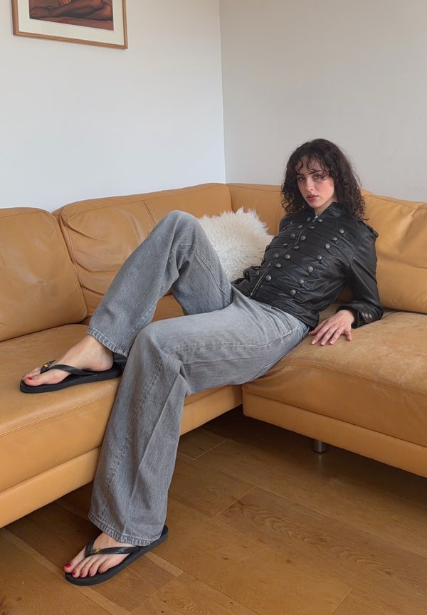 Kvinne med krøllete hår som lener seg tilbake på en lysebrun skinnsofa, iført svart jakke med knapper, grå vide jeans og svarte flip-flops på tregulvet.