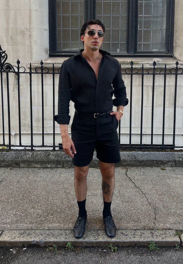 Mann trägt schwarzes Hemd mit Knöpfen, schwarze Shorts, schwarze Schuhe und Sonnenbrille, steht auf dem Bürgersteig neben einem schwarzen Metallzaun und einem Steingebäude.