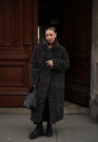 Mujer con un abrigo largo negro de textura rizada, botas negras y pendientes dorados, sosteniendo un bolso negro, de pie frente a puertas de madera.