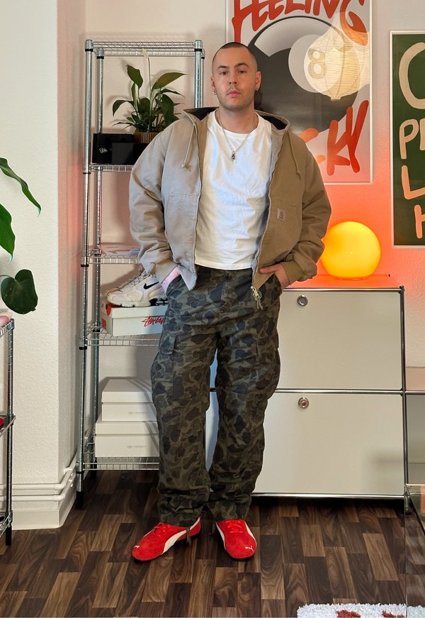 Giacca con cappuccio beige, t-shirt bianca, pantaloni cargo mimetici e scarpe da ginnastica rosse. Stanza con piante, mensole e luci decorative.
