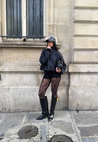 Ung kvinna i svart plisserad jacka, svarta spetsstrumpbyxor, stövlar med hög klack och keps, lutande mot en stenbyggnadsvägg på en stadsgata.