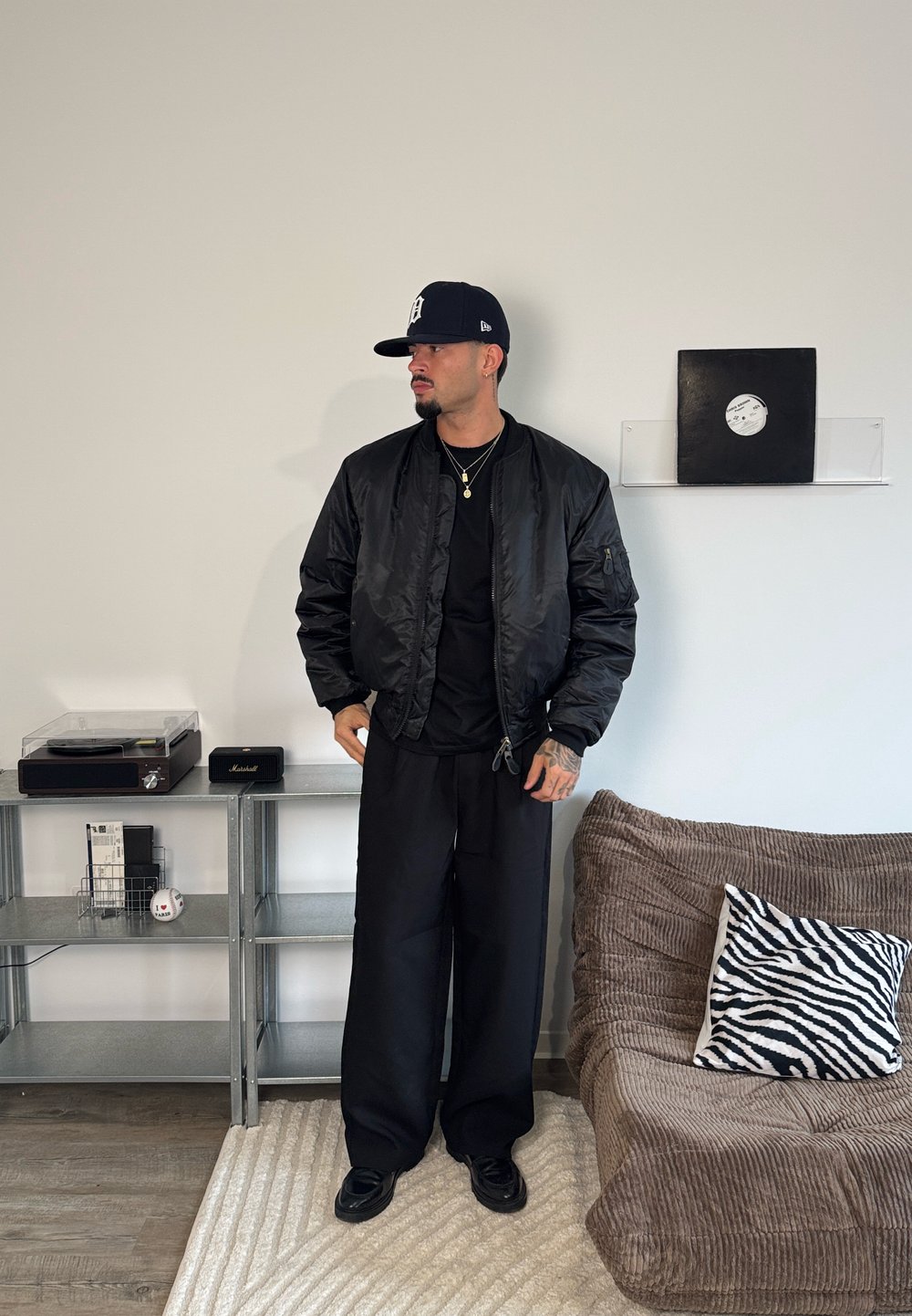 Cazadora bomber negra con una textura suave y brillante, llevada sobre una camiseta negra. Pantalones negros sueltos y una gorra azul marino. Los accesorios incluyen un collar dorado.