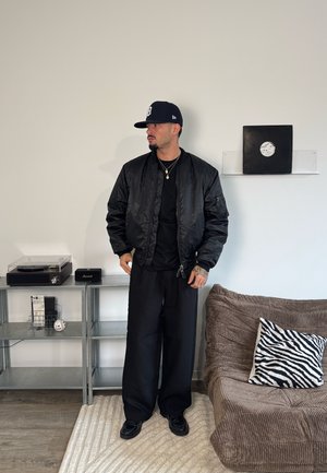 Cazadora bomber negra con una textura suave y brillante, llevada sobre una camiseta negra. Pantalones negros sueltos y una gorra azul marino. Los accesorios incluyen un collar dorado.