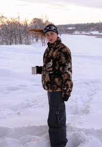 Fleecevest in bruin-zwart patroon, met ritssluiting aan de voorkant en een buidelzak; gecombineerd met zwarte cargobroek en een zilveren mok vasthoudend.