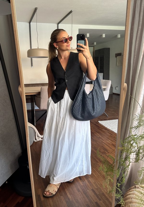 Chaleco negro con botones, falda blanca fluida, sandalias marrones de tipo deslizante, bolso de mimbre azul y gafas de sol rojas y redondas. Un espejo refleja un interior moderno.