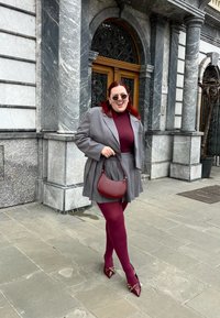 Γκρίζα σακάκι και πλισέ σορτς πάνω από μπορντό τοπ και καλσόν, συνδυασμένα με μπορντό τσάντα και κόκκινες μυτερές γόβες, μπροστά από μαρμάρινη πρόσοψη.