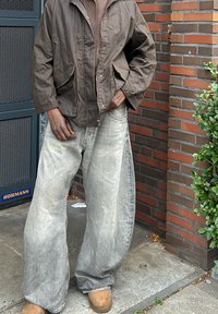 Braune Zip-Jacke, braunes Langarmshirt, ausgewaschene weit geschnittene Jeans und hellbraune Stiefel. Dunkel gemusterte Handschuhe und strukturierte Stoffdetails.