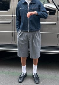 Denim zip-up jacket with two chest pockets, grey pleated shorts, white crew socks, and black loafers. Background features a vehicle.