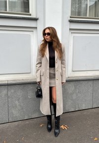 Abrigo a rayas beige con botones dorados, combinado con una blusa negra transparente y una falda corta. Botas negras hasta la rodilla y un pequeño bolso negro.