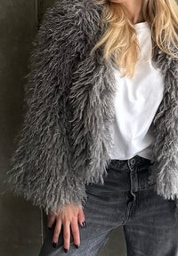 Veste en fausse fourrure grise à texture shaggy, avec manches longues et coupe décontractée, portée sur un t-shirt blanc et un jean gris.