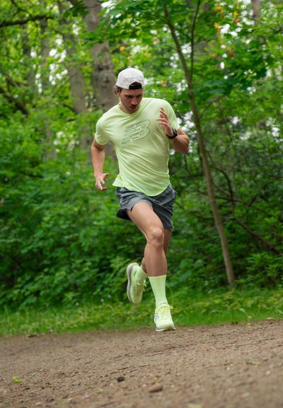 Hombre con ropa deportiva amarilla y gorra blanca corriendo por un camino de tierra en un bosque verde, con aspecto concentrado y a mitad de zancada.