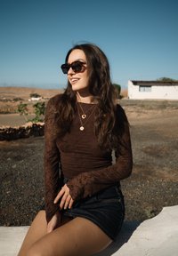 Γυναίκα με μακριά καστανά μαλλιά, φοράει γυαλιά ηλίου, μακρυμάνικη μπλούζα σκούρου καφέ χρώματος, τζιν σορτς και χρυσά κολιέ, κάθεται σε εξωτερικό χώρο σε ηλιόλουστο αγροτικό περιβάλλον.