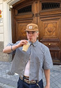 Kratke rokave s karo vzorcem v modri in beli barvi, nosi čez belo majico brez rokavov. Bež kapa z logotipom, pije ledeno kavo iz plastične skodelice.