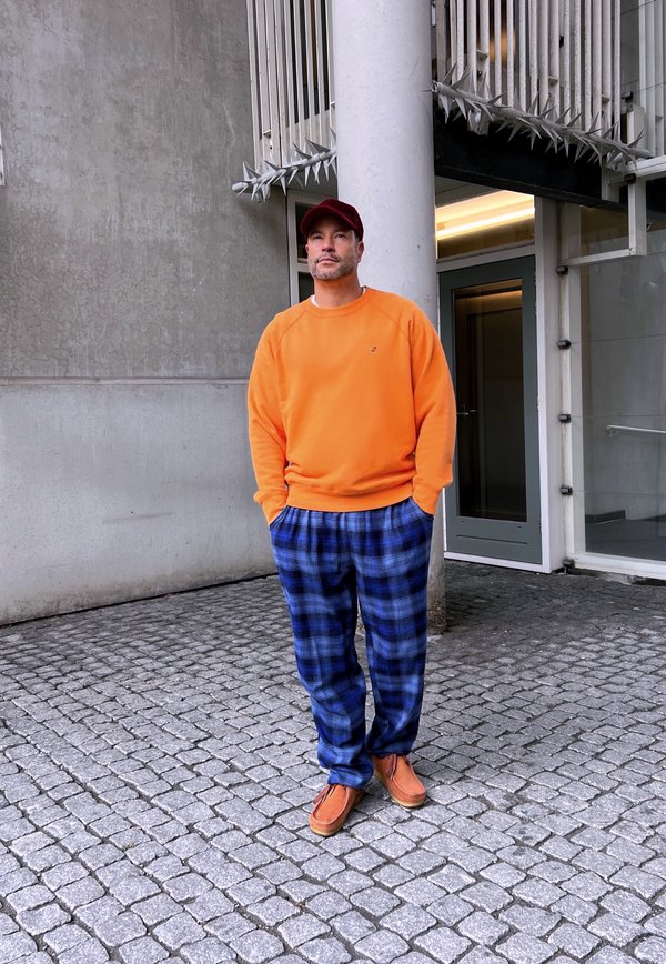 Sweatshirt laranja, calças de pijama xadrez azuis e pretas, e sapatos mocassim castanhos. De pé contra uma parede cinza e um pilar.
